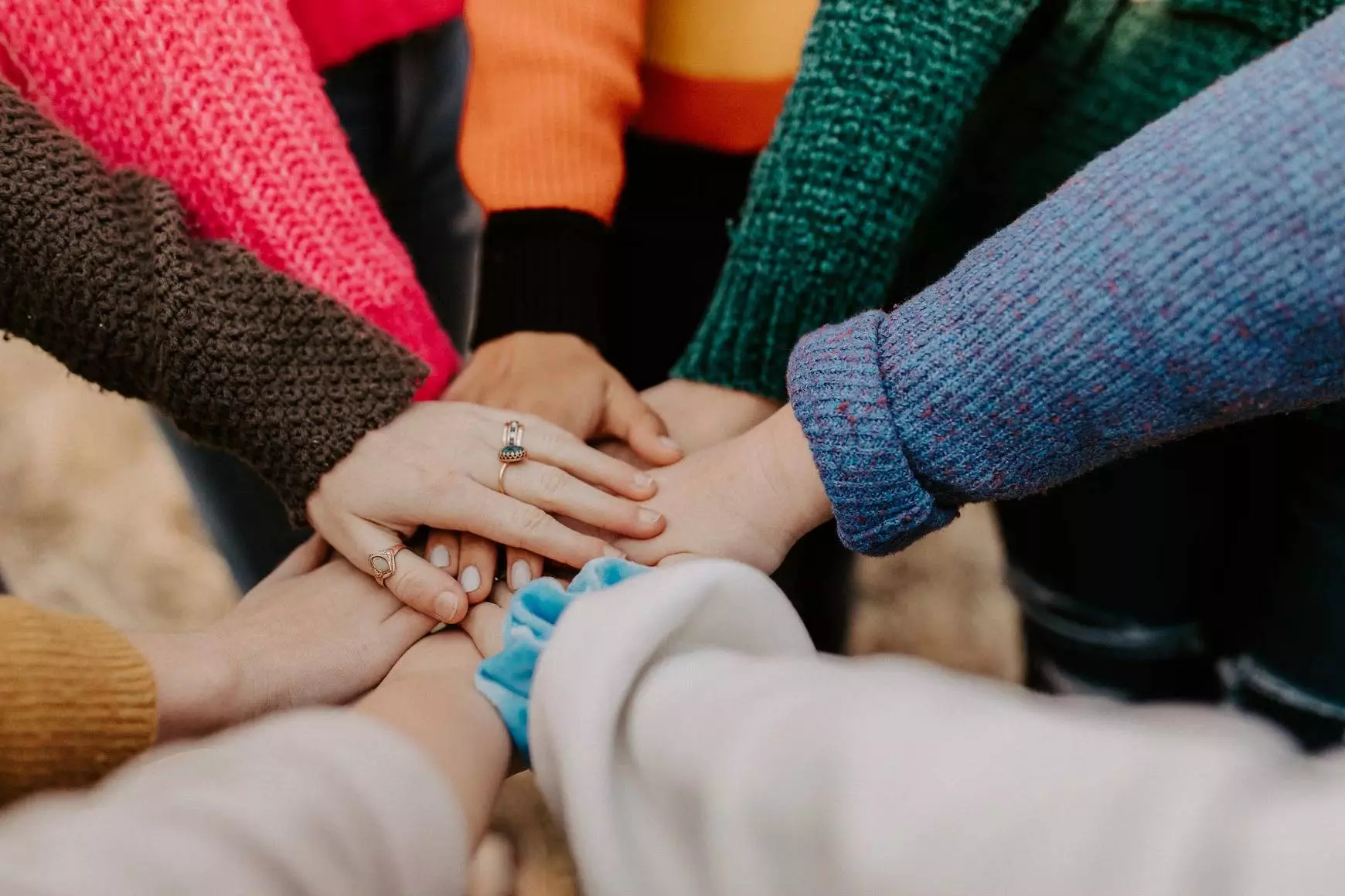 A close-up of several hands holding each other. The hands are all wearing rings and the background appears to be a dark, blurry orange sweater. There is one person in the center with their arm visible, while the others have their arms hidden behind them.