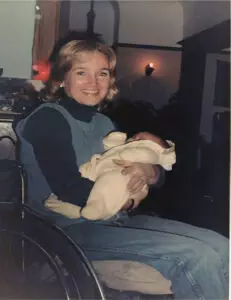 Shelley with a joyful expression, wearing a blue turtleneck and vest, seated in a wheelchair, holding a baby wrapped in a white blanket, in a dimly lit room with a candle in the background.
