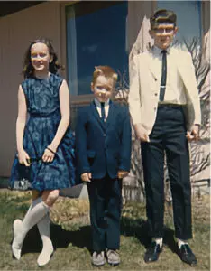 The image shows three children outside on a sunny day: a girl, Shelley, in a blue floral dress with white knee-high socks, a young boy in a dark suit with red hair, and an older boy in a white shirt and dark pants, all smiling. The vintage-style photo hints at a mid-20th-century setting in a residential area.