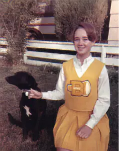 Shelley in a white blouse and yellow cheerleading outfit with a letter 'B' stands next to a black dog in a sunny yard with a white picket fence and camper in the background.